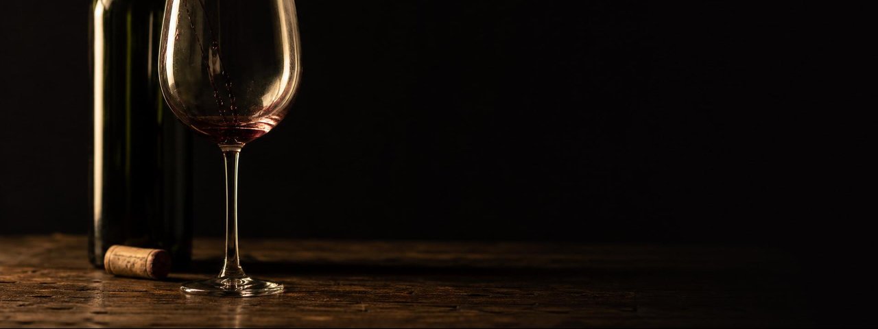 An empty wine glass with the last traces of red wine, a label-less bottle and a cork on a dark table.