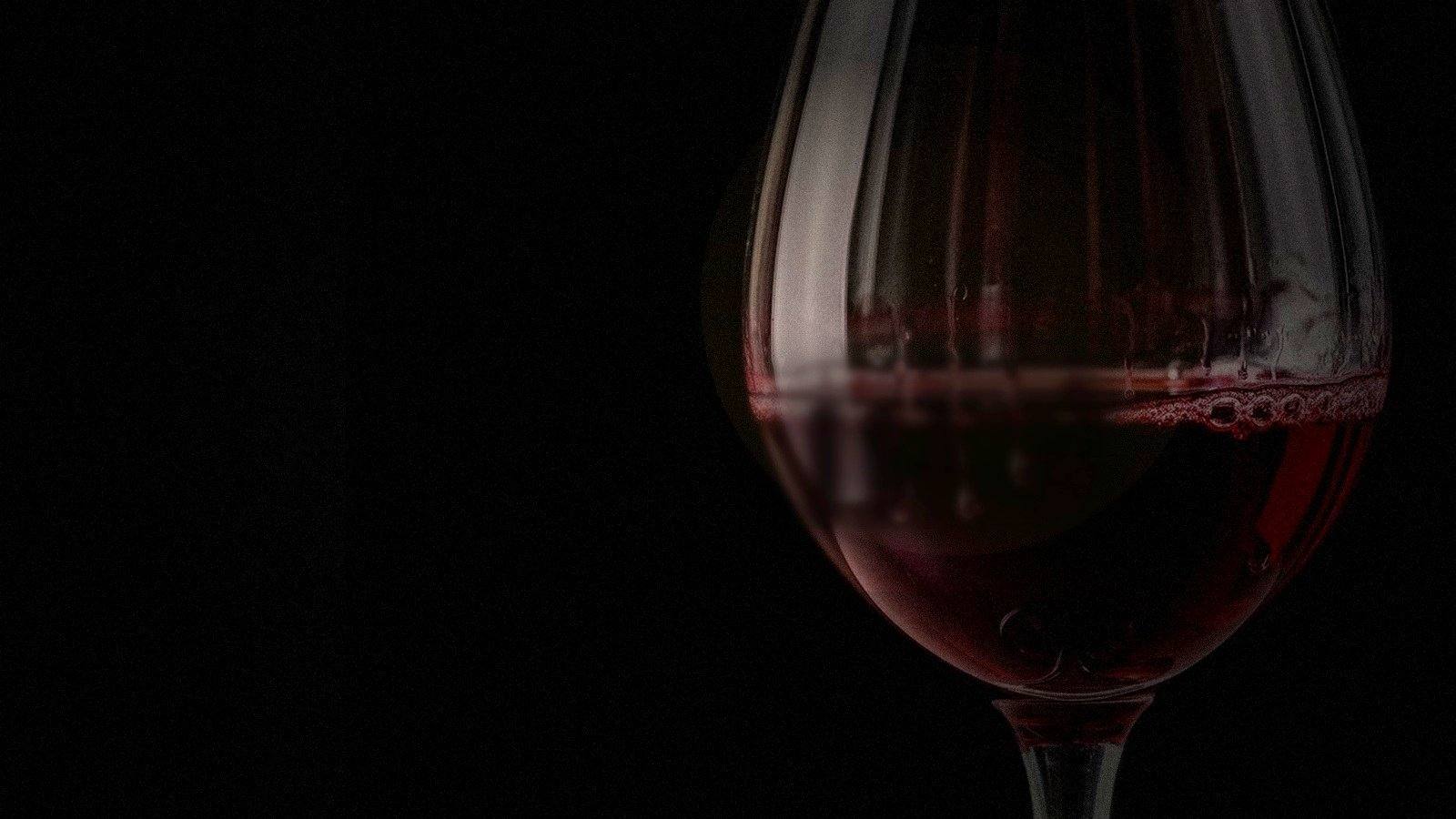 A glass of dark red wine in deep shadow, lit by a faint warm rim of light.