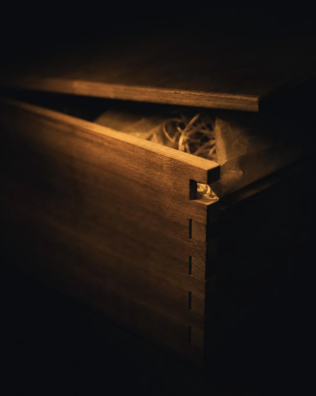 An original wooden case with its lid ajar, wood-wool inside, dovetail joints in raking light.