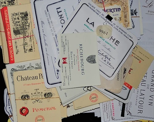 A pile of loose printed wine labels imitating those of well-known Burgundy and Bordeaux estates, with a vintage stamp among them.