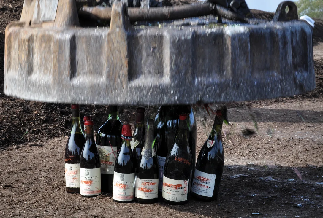 A row of old bottles stamped COUNTERFEIT in red, standing on bare earth beneath the raised plate of an industrial magnet; the labels carry the names of Burgundy and Champagne houses.